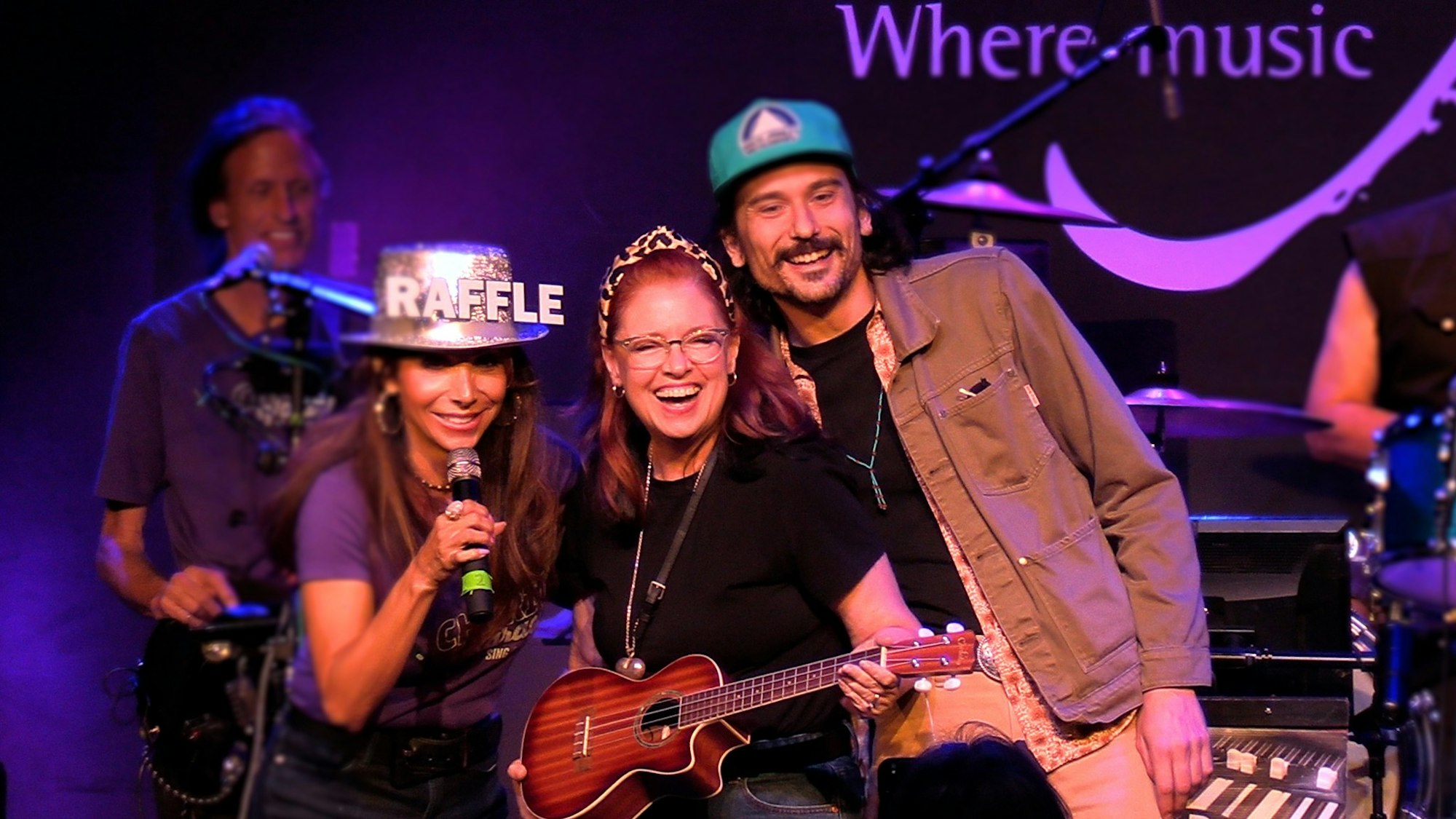 A group of people on stage, one holding a microphone and wearing a "RAFFLE" hat. Another person is holding a ukulele.