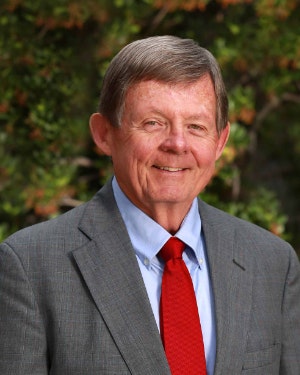 An older man in a suit and red tie, smiling outdoors with a blurred green background.