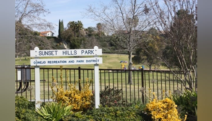 Sign for Sunset Hills Park, Conejo Recreation and Park District, with trees, grassy field, and play area in the background.