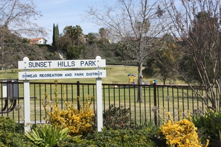 Sign for Sunset Hills Park, Conejo Recreation and Park District, with trees, grassy field, and play area in the background.