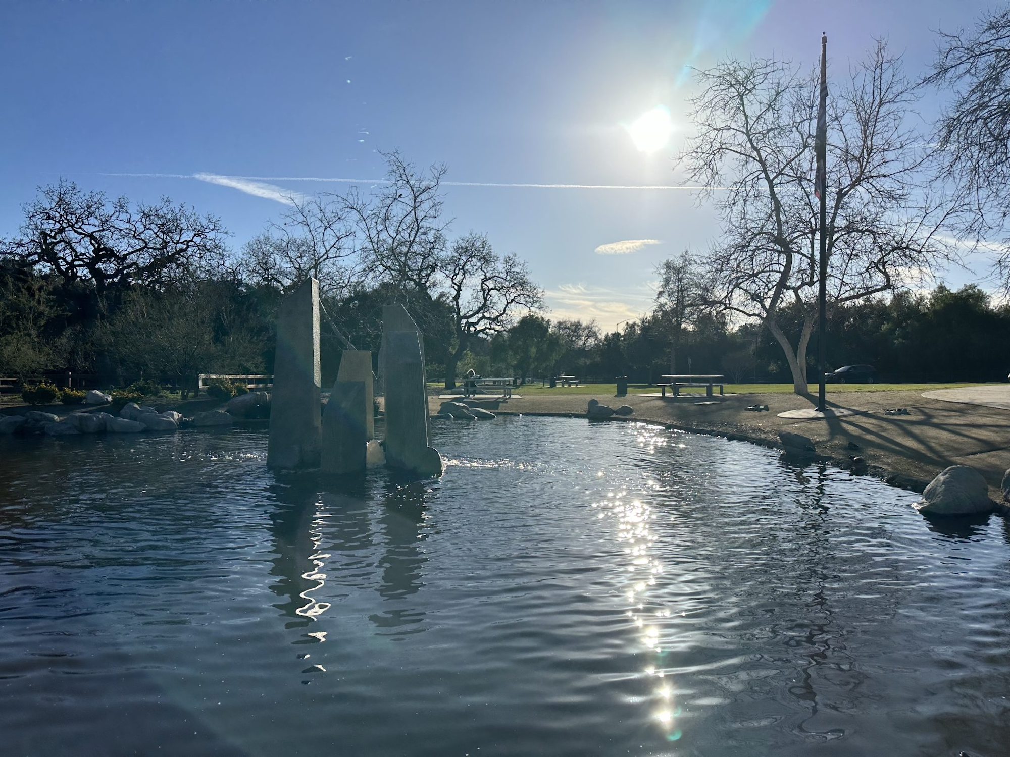 A pond with stone structures, reflections on water, bare trees, sunny sky, and a park setting.
