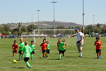 Kids in green and orange shirts are playing soccer on a field with a coach nearby.