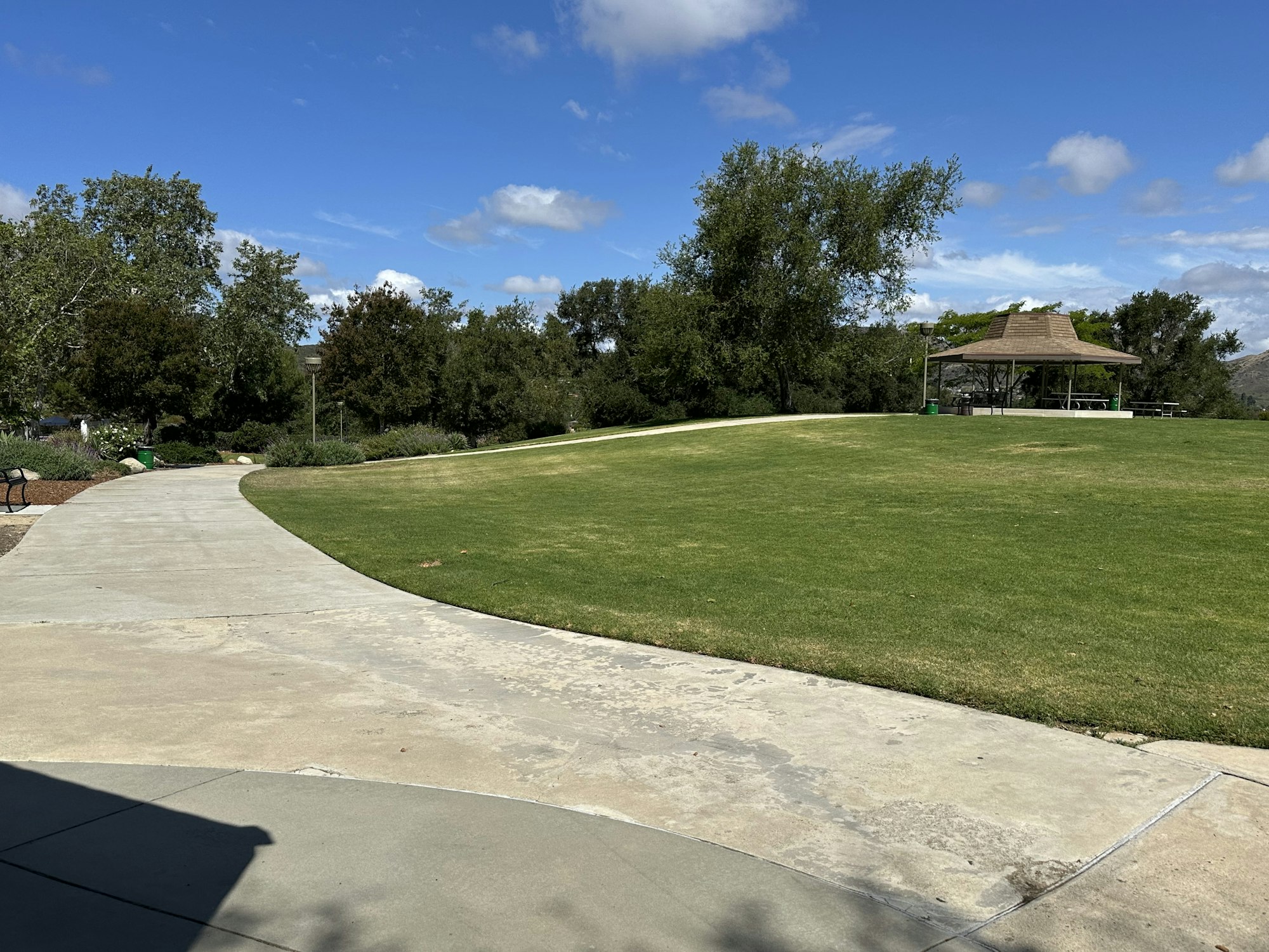 A scenic park view with a winding path, grassy area, trees, and a gazebo under a blue sky with clouds.