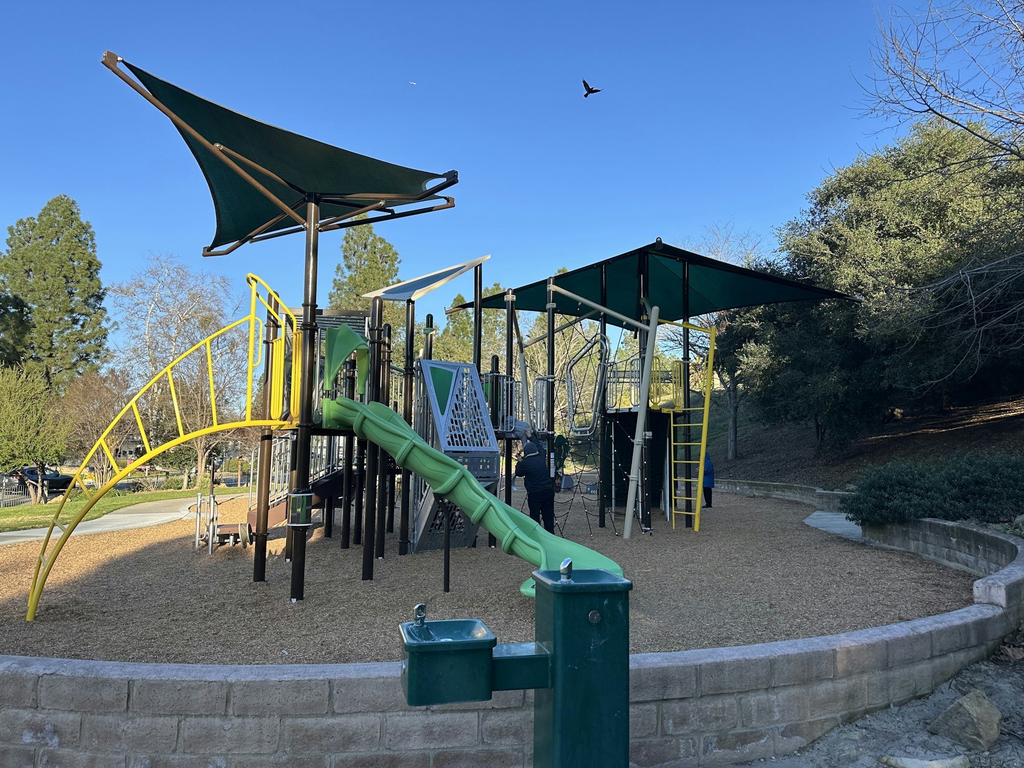Playground with slides, climbing structures, and shade. A drinking fountain is in the foreground, with trees and a bird in the background.