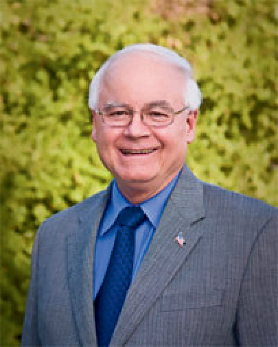 An older man in a suit smiles in front of a leafy background.