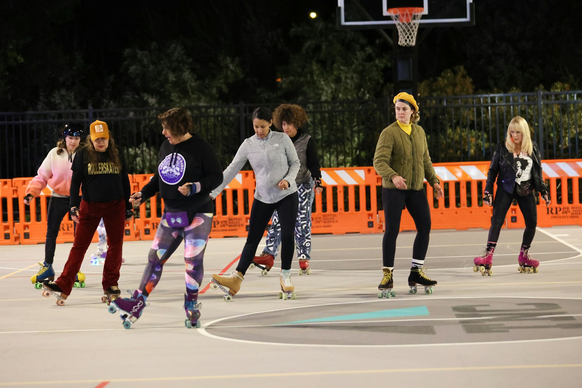 A group of people roller skating together at night on a court with an orange barrier in the background.
