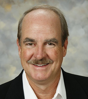 A man with a mustache smiles, wearing a black blazer and white shirt, against a neutral background.