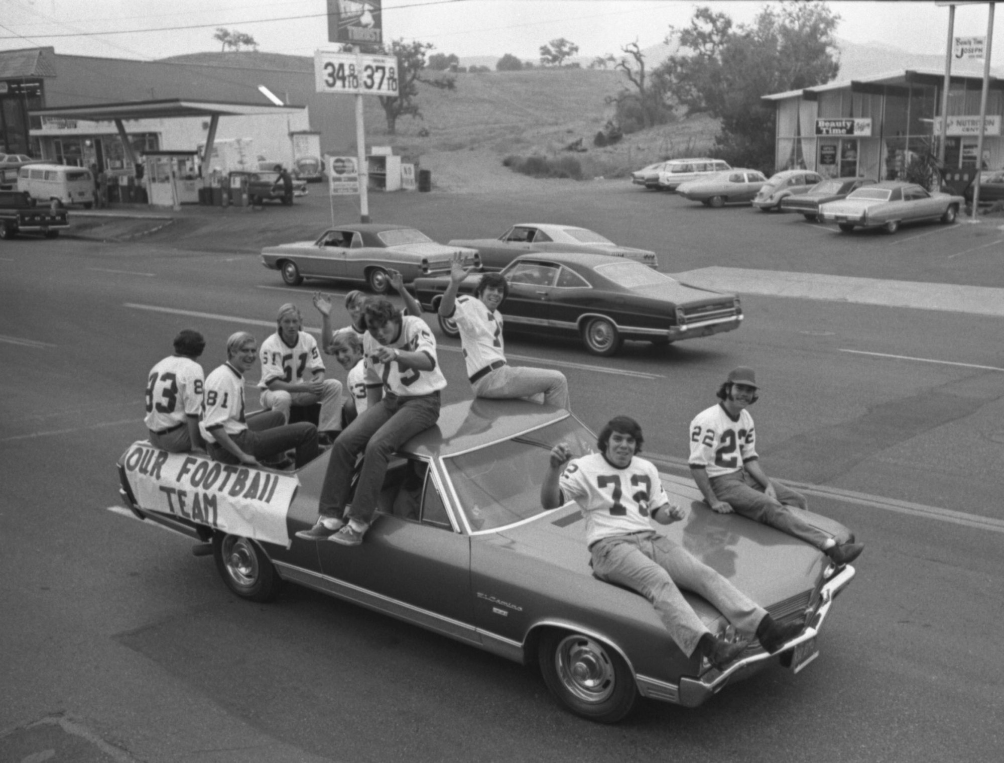 Football team on a car for a parade. They're wearing jerseys and waving. Retro street scene with old cars and gas price signs.