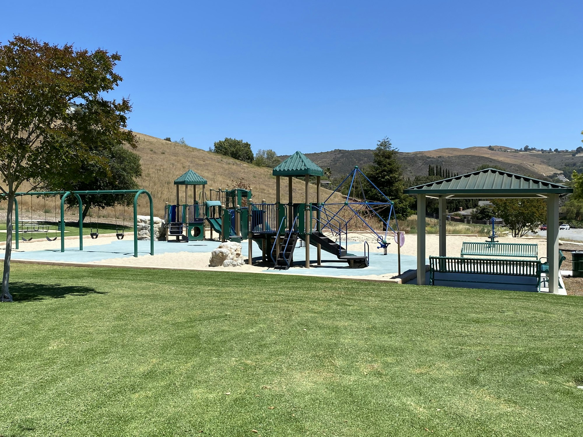 Playground with swings, climbing structures, and benches on a sunny day. Surrounded by grass, trees, and hills.