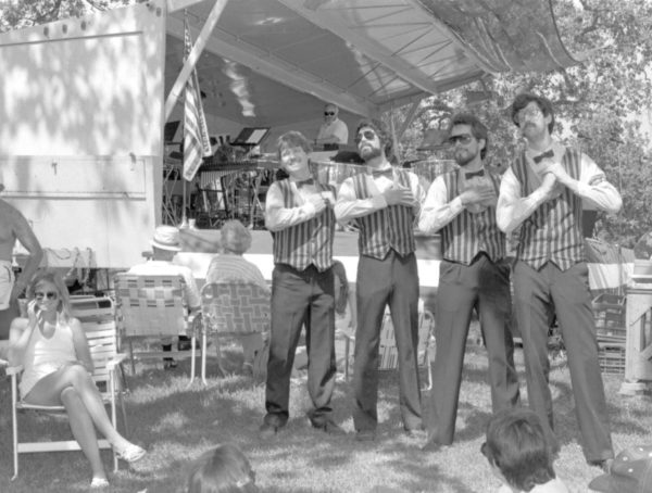 Four men in matching vests and bow ties perform outdoors, with people seated nearby and a stage in the background.