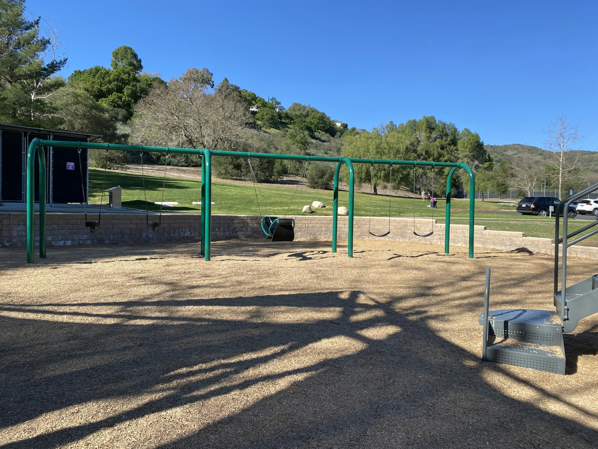 A playground with swings, surrounded by grass and trees, under a clear blue sky.