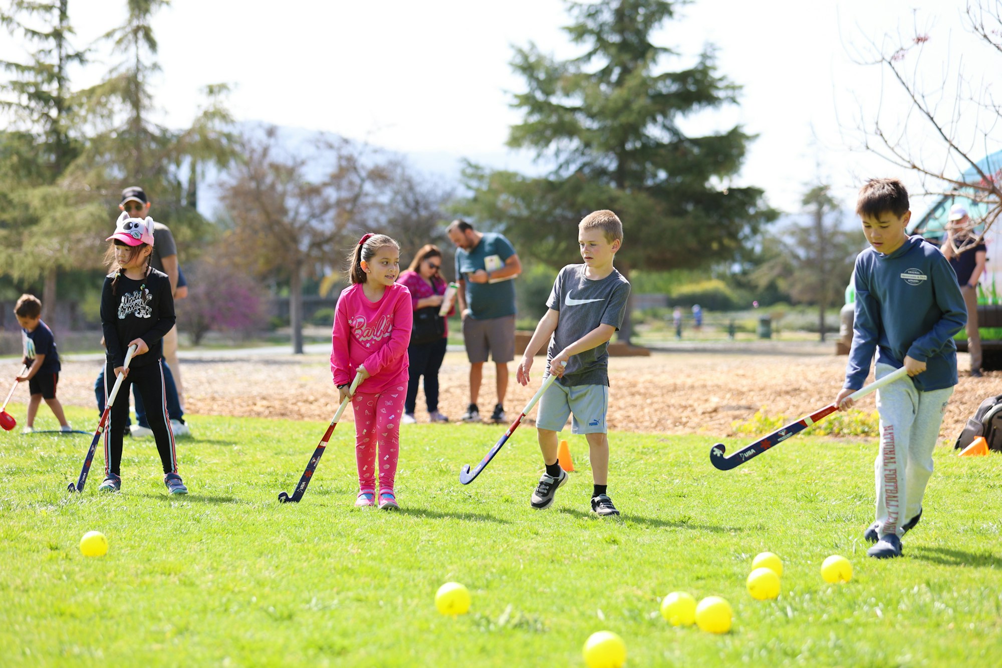 Kids playing field hockey on grass while adults watch nearby in a park setting.