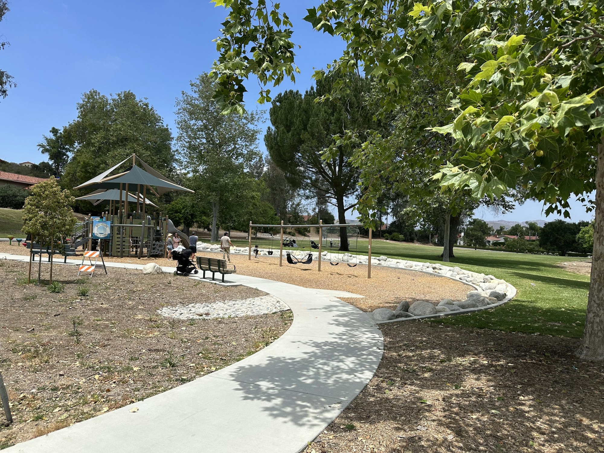 A sunny park scene featuring a playground, walking path, benches, and people enjoying the outdoors among trees.