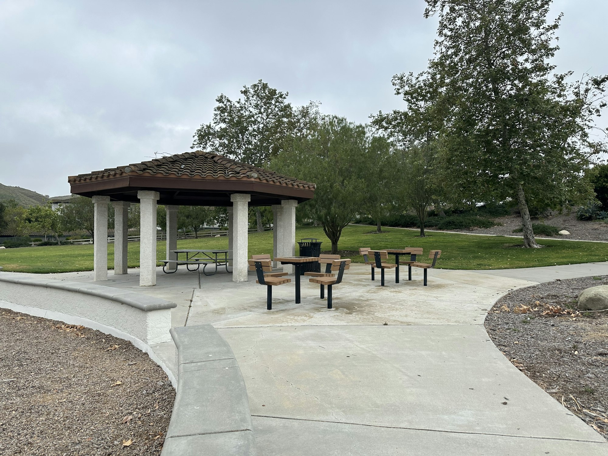 A park setting featuring a gazebo and picnic tables surrounded by trees and grass, on a cloudy day.