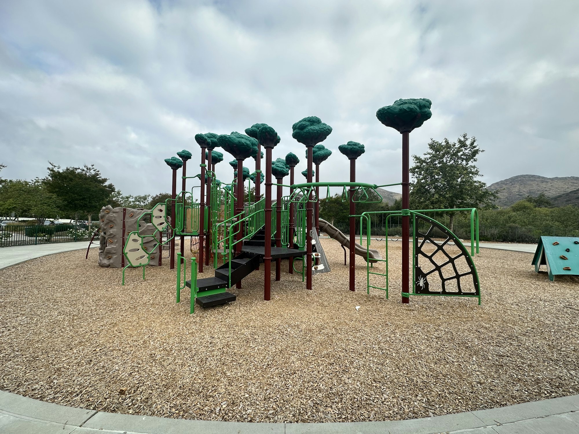A colorful playground with tree-like structures, climbing walls, slides, and a rubber mulch surface, set against a cloudy sky.