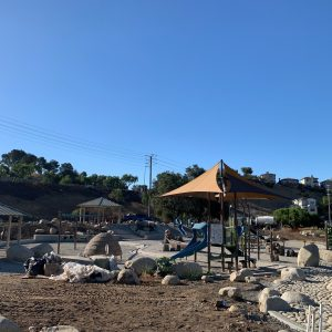 A playground with slides, shade structures, and rocky terrain under a clear blue sky.