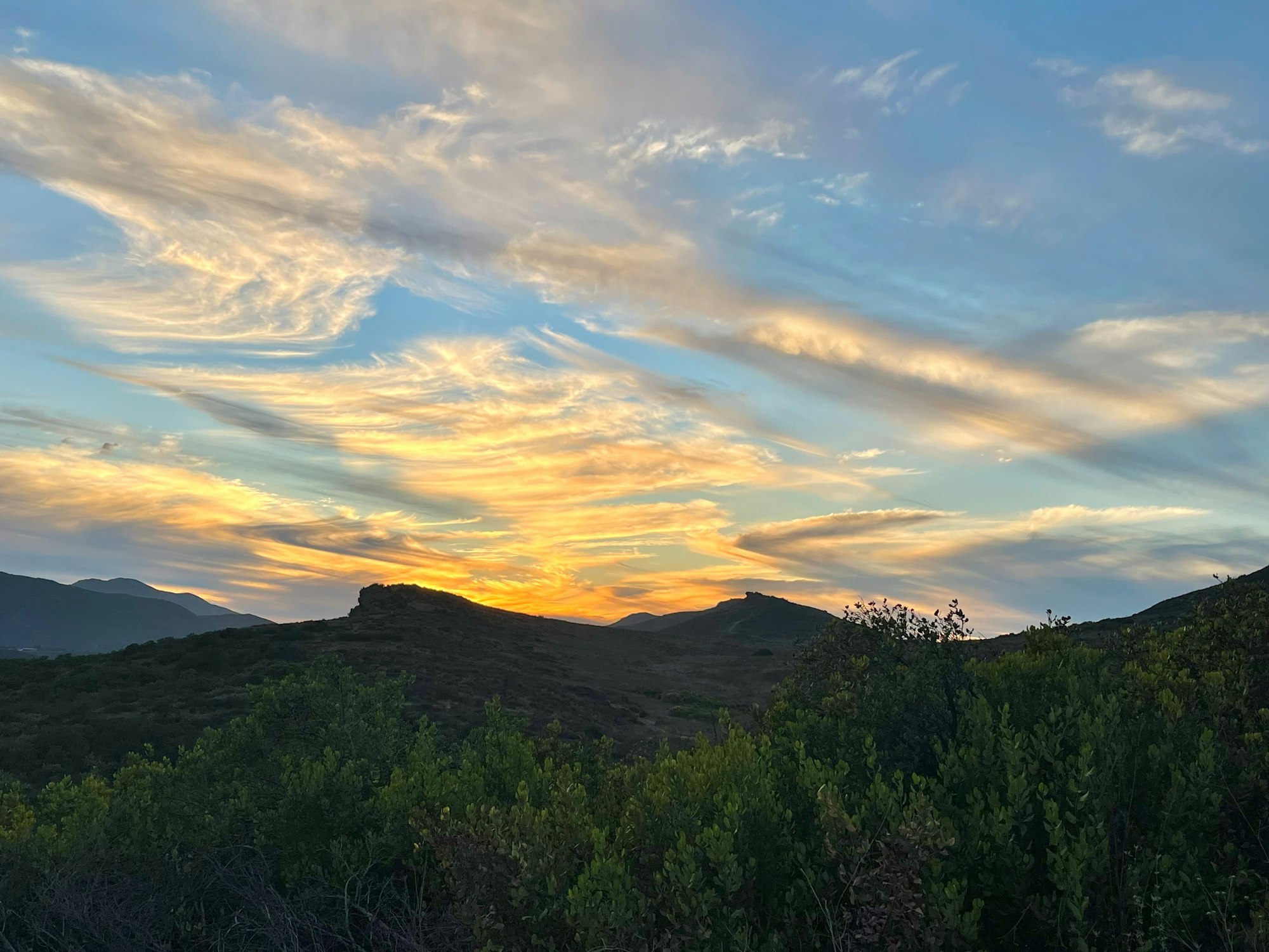 A scenic sunset over hills with colorful clouds and lush green foliage in the foreground.