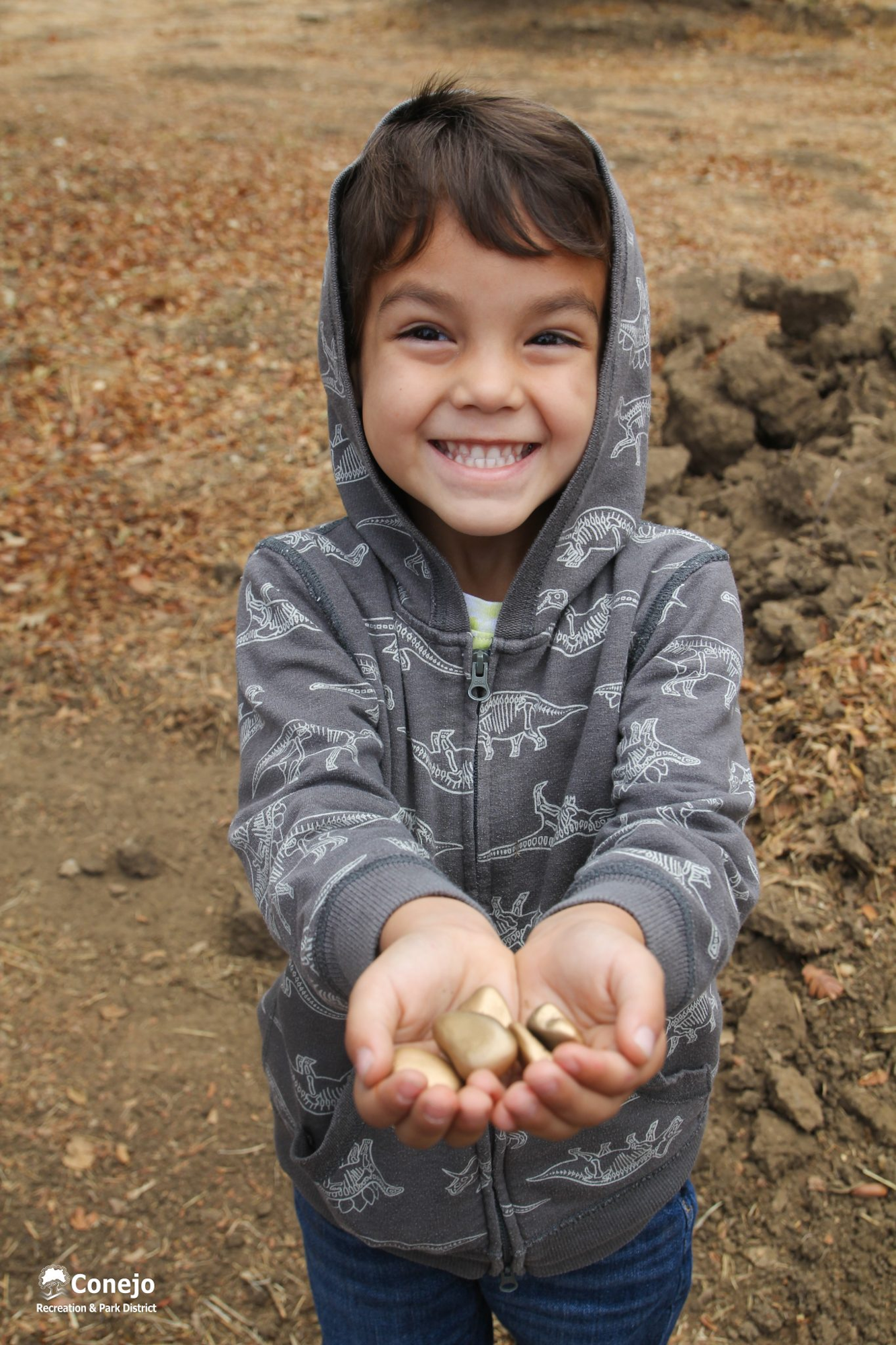 A smiling child in a dinosaur hoodie holding rocks outdoors.