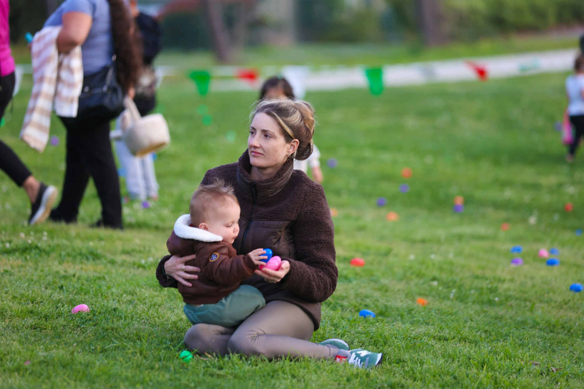 A woman sits on grass holding a baby with colorful eggs around them. Other people are in the background.