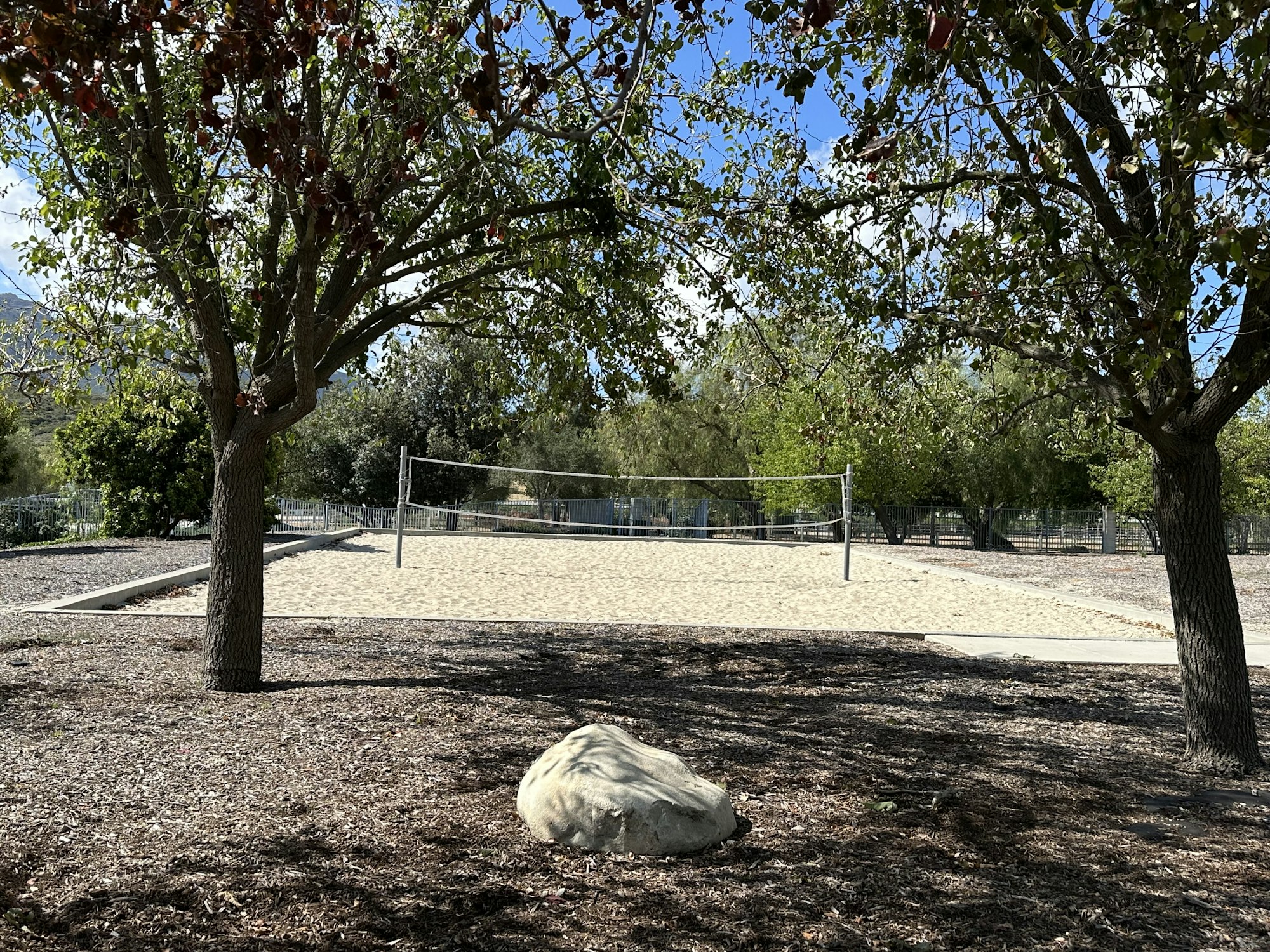 A sandy volleyball court surrounded by trees and greenery, with a large rock in the foreground.