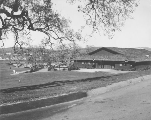 A large wooden building surrounded by trees and grass, with a paved road in the foreground.