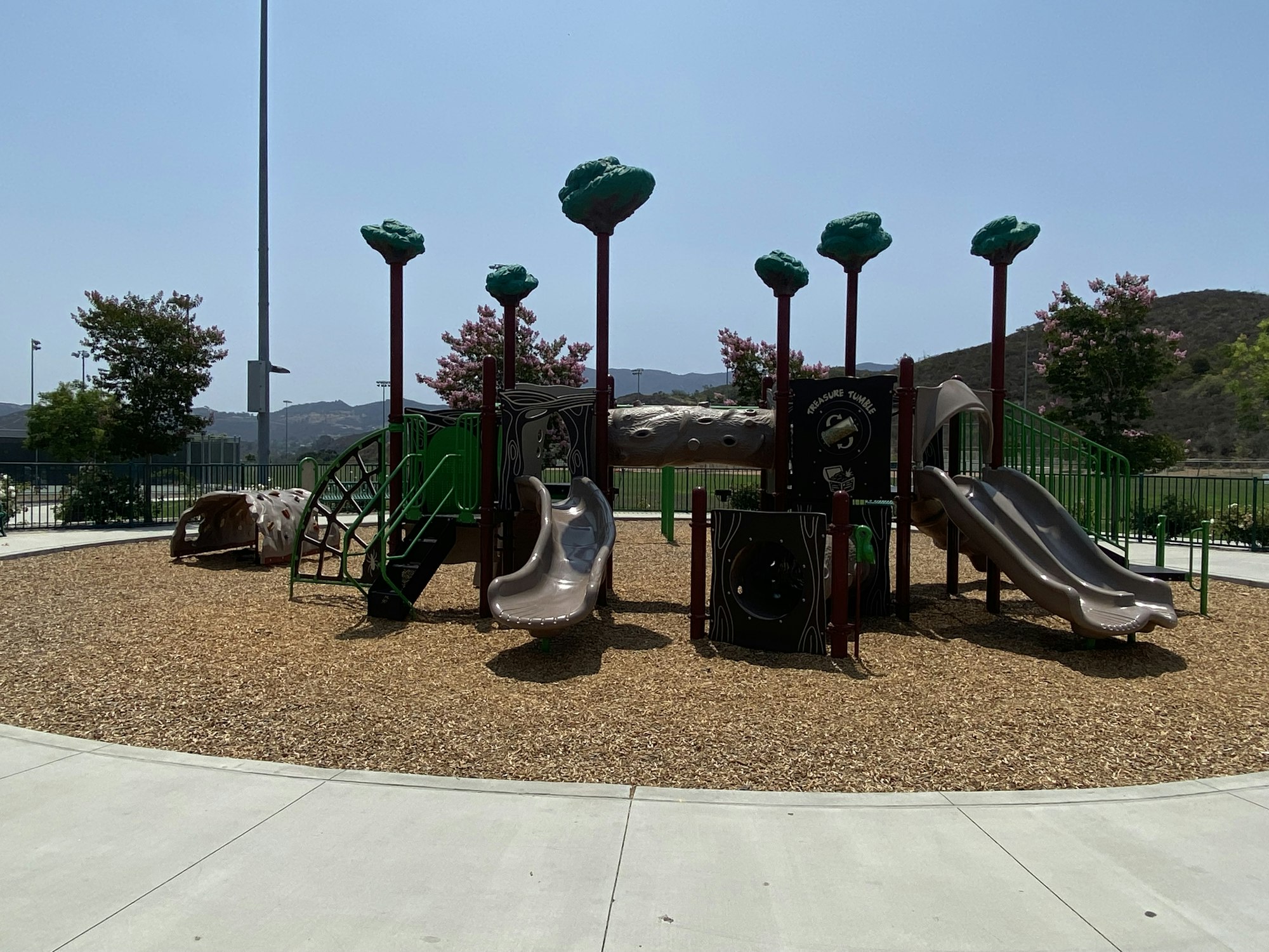 Children's playground with slides, climbing structures, and artificial trees on woodchip ground.
