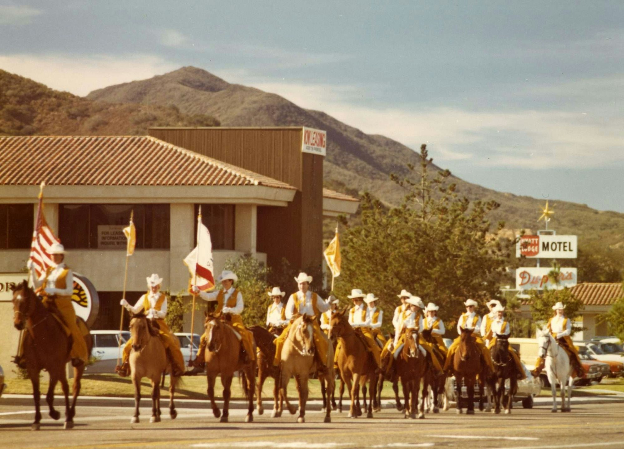 A group of riders on horseback in a parade, holding flags, with buildings and hills in the background.