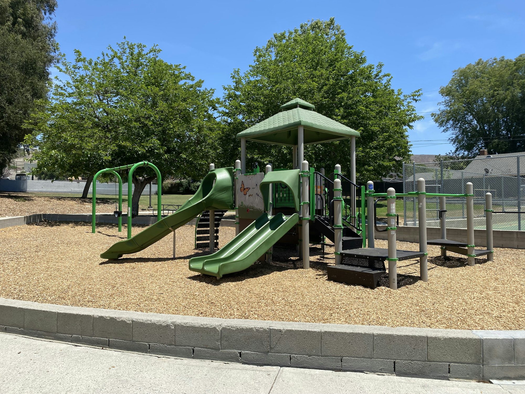 Playground with green slides, climbing structure, and woodchip ground cover, surrounded by trees under a clear blue sky.
