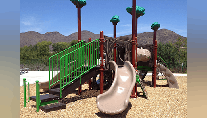 Playground with slides, climbing structure, green railings, and tree-themed decorations, set against a mountain backdrop.