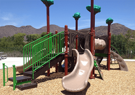 Playground with slides, climbing structure, green railings, and tree-themed decorations, set against a mountain backdrop.