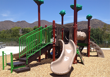 Playground with slides, climbing structure, green railings, and tree-themed decorations, set against a mountain backdrop.