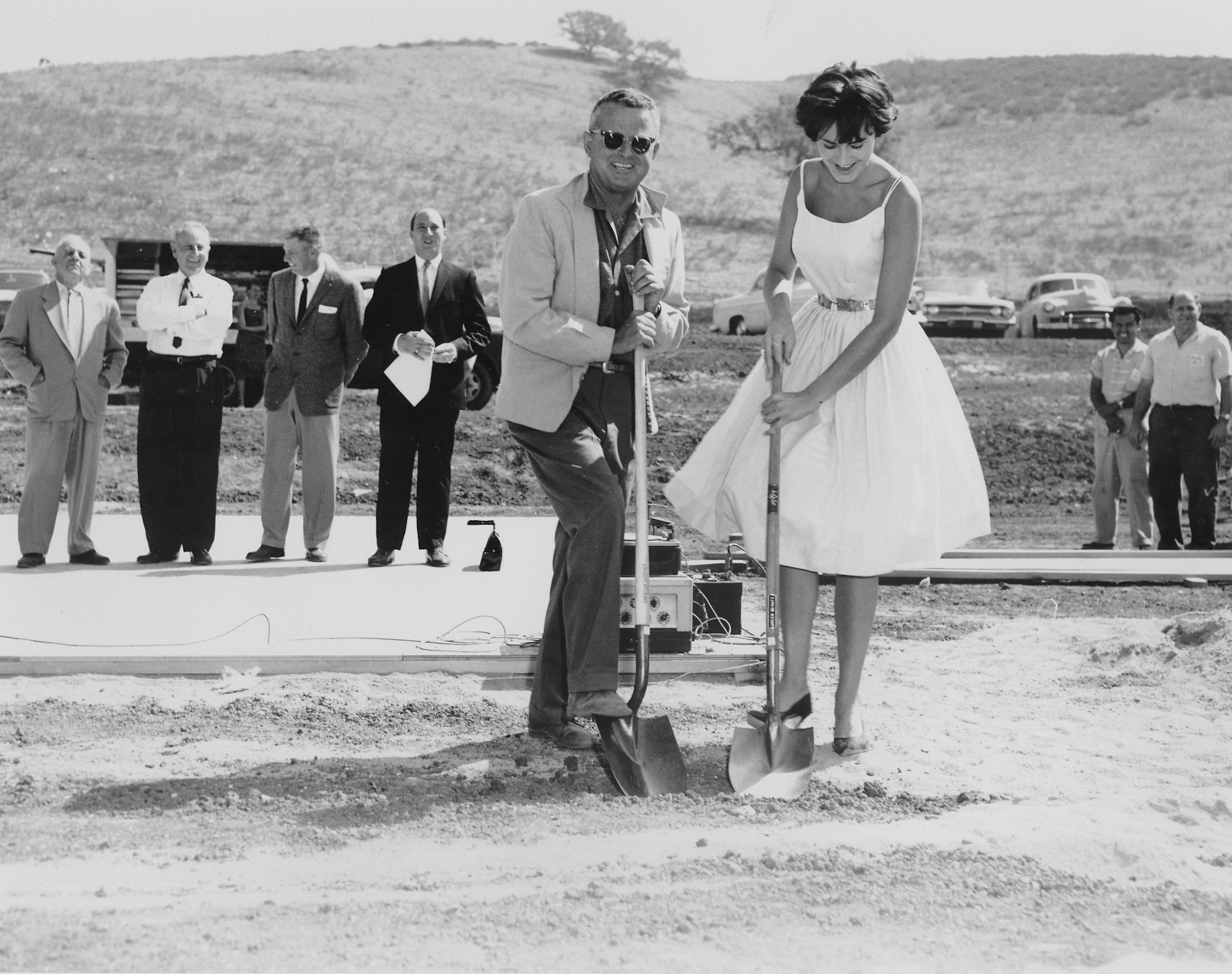 A groundbreaking ceremony with a man and woman using shovels, surrounded by several onlookers and cars in the background.
