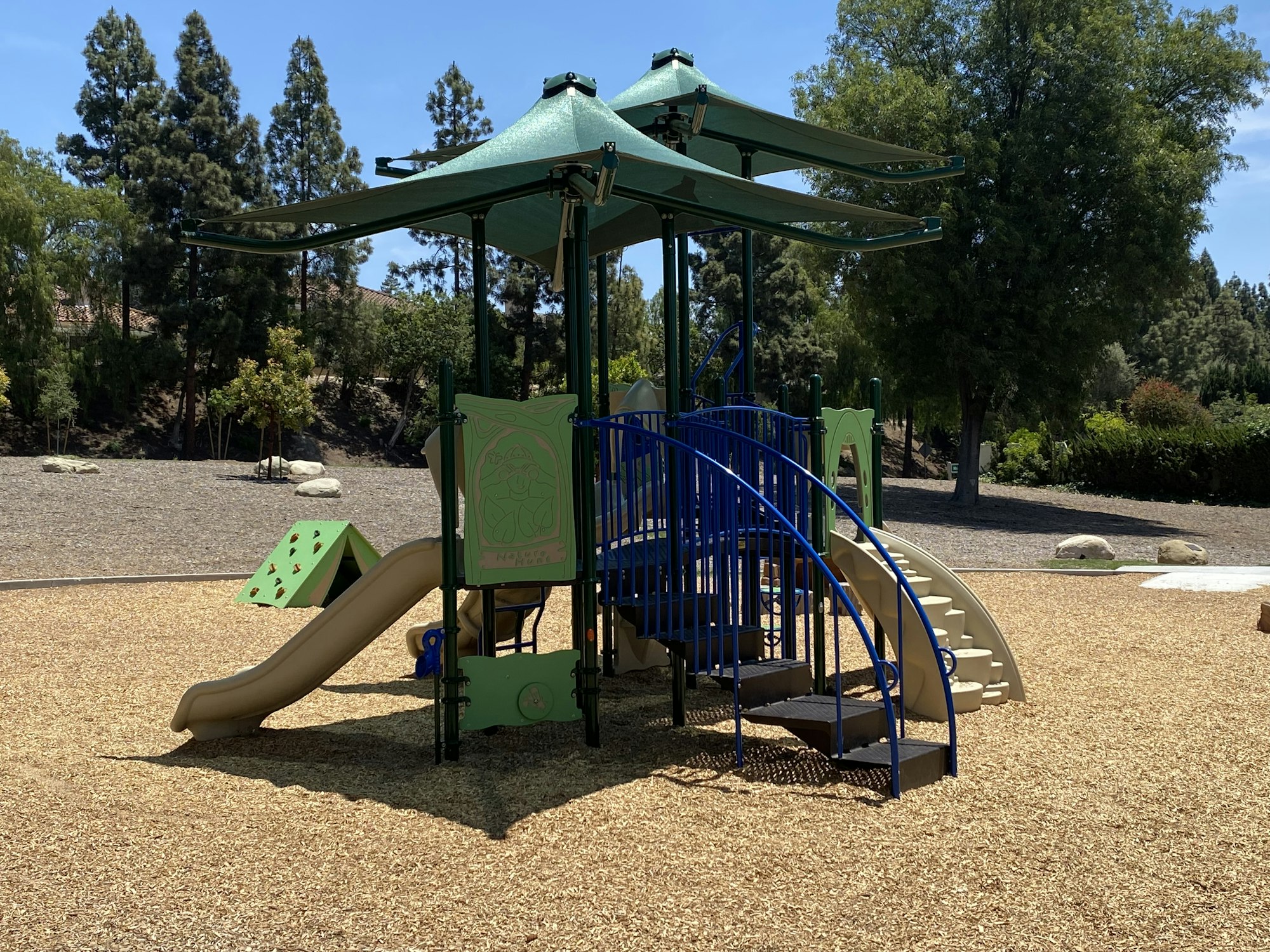 A playground with a slide, climbing structure, and steps surrounded by wood chips and trees.