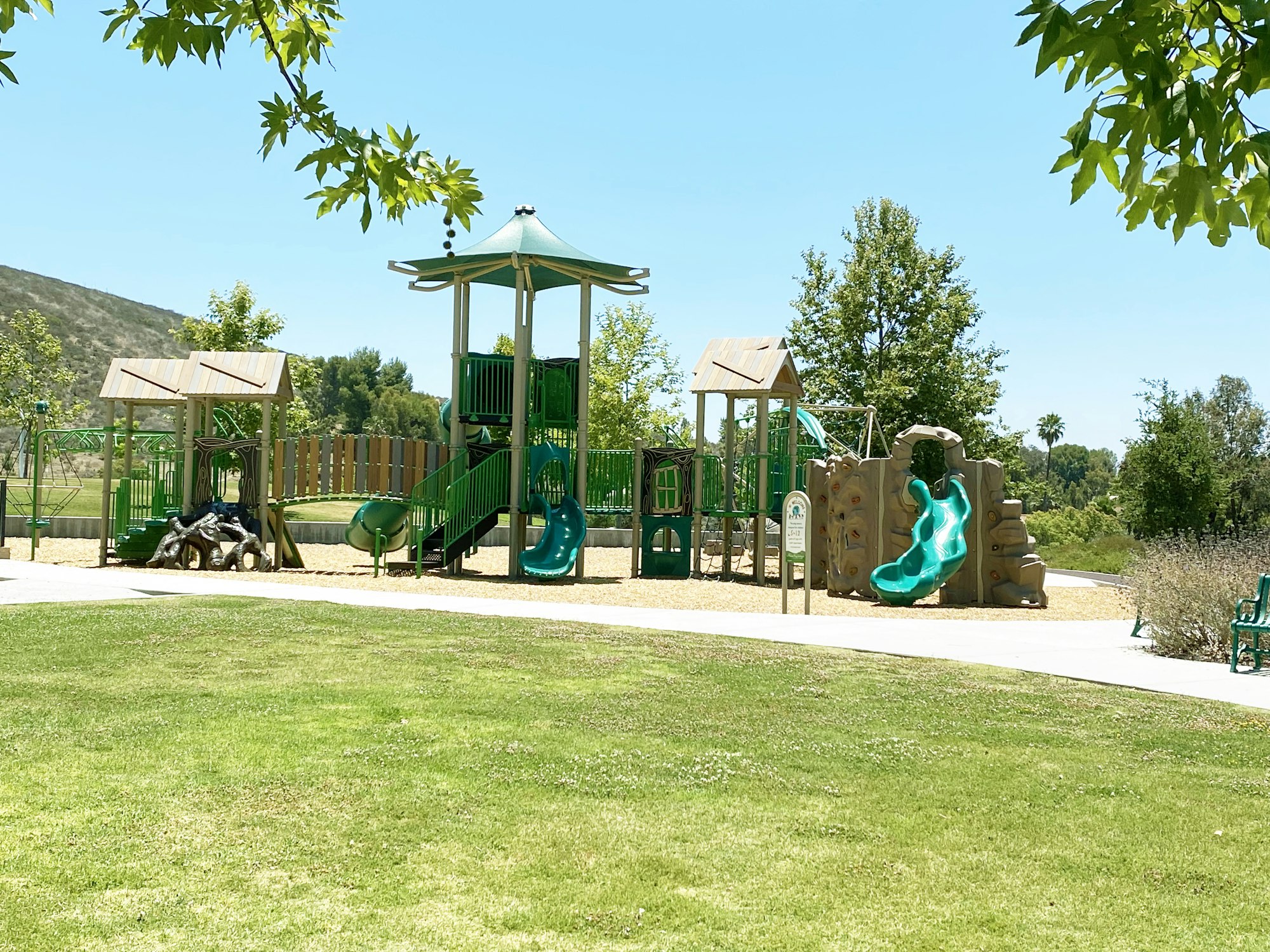 Playground with slides, towers, and climbing structures surrounded by grass and trees under a clear blue sky.