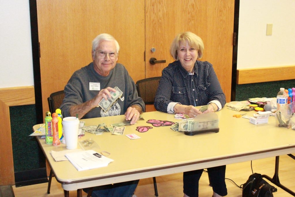 Two people sit at a table with cash, paper items, and drinks, appearing to be organizing or counting money.