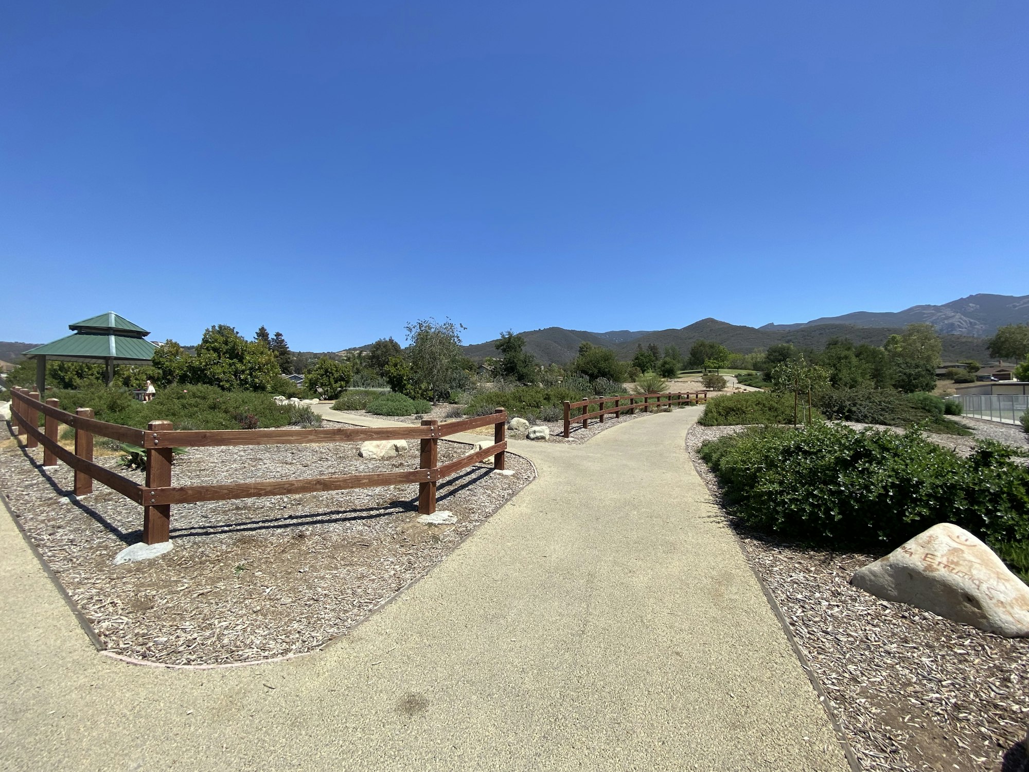A paved pathway with wooden fences, greenery, a gazebo, and mountains in the background under a clear blue sky.