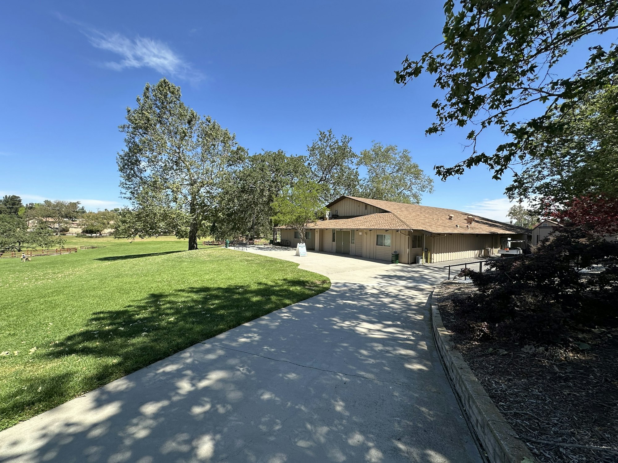 A sunny park scene featuring a pathway, green grass, trees, and a building. Clear blue sky in the background.