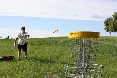 Person playing disc golf, throwing a frisbee towards a metal basket in a grassy field.