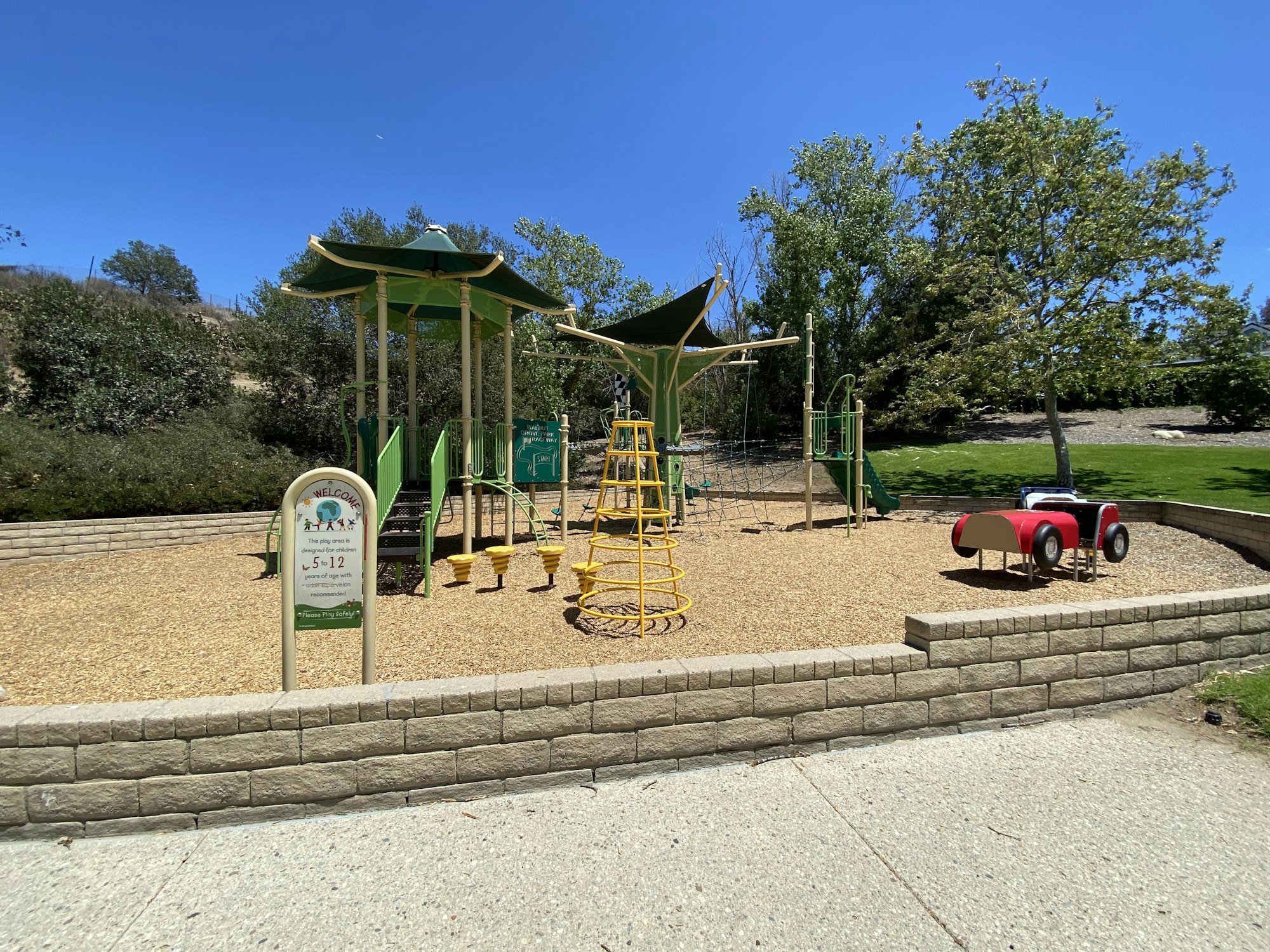 Children's playground with slides, climbing structures, and a toy car. Surrounded by greenery under a clear blue sky.