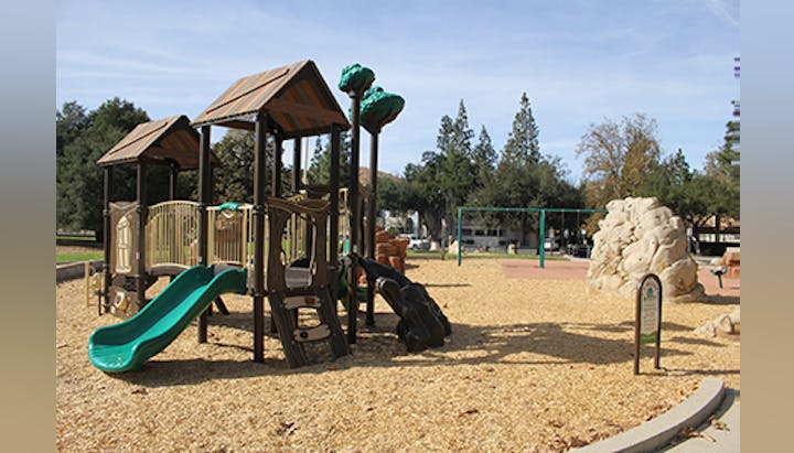 A playground with slides, climbing structures, swings, and a rock wall on wood chips, surrounded by trees.