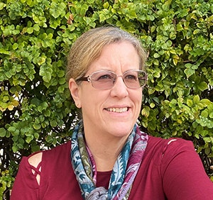 Person wearing glasses and a scarf, sitting in front of green foliage, smiling.