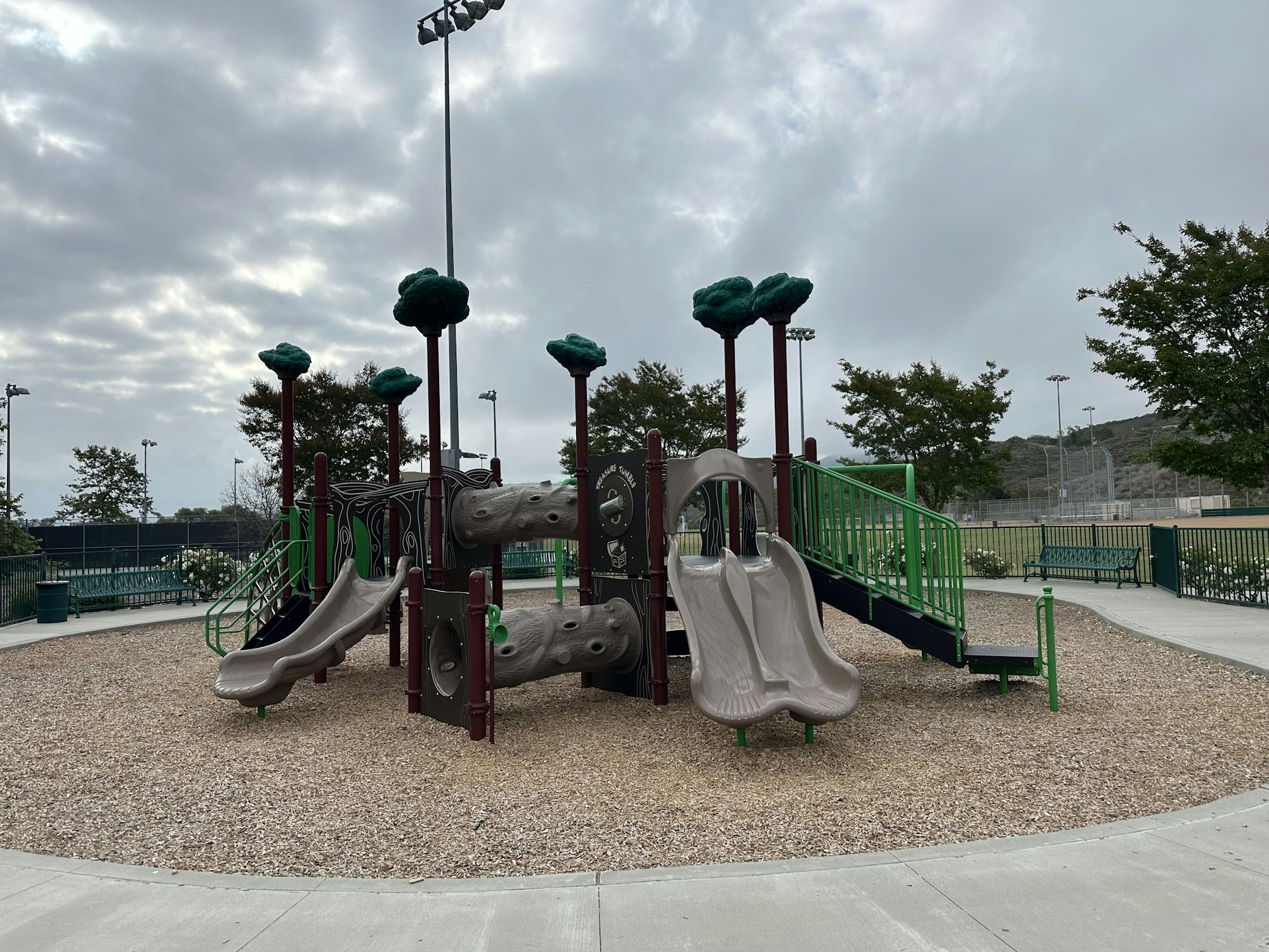 A playground with two slides, climbing structures, and green features, set in a gravel area under an overcast sky.