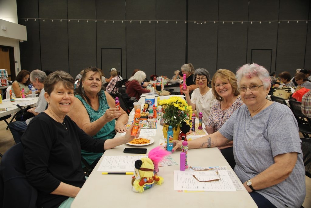 A group of women sitting at a table with bingo cards, daubers, and snacks, smiling at the camera.