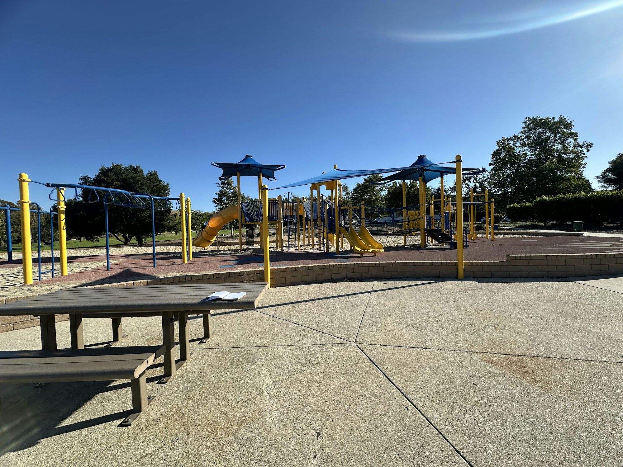 A playground with yellow slides and climbing structures, surrounded by a grassy area and clear blue sky. A picnic table is nearby.