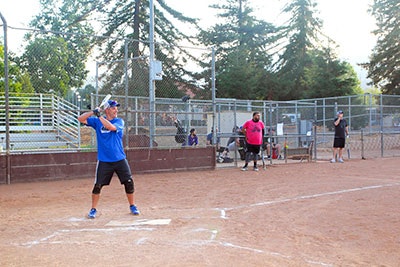 A person in a blue shirt is preparing to bat in a softball game on a dirt field with a backstop and onlookers.