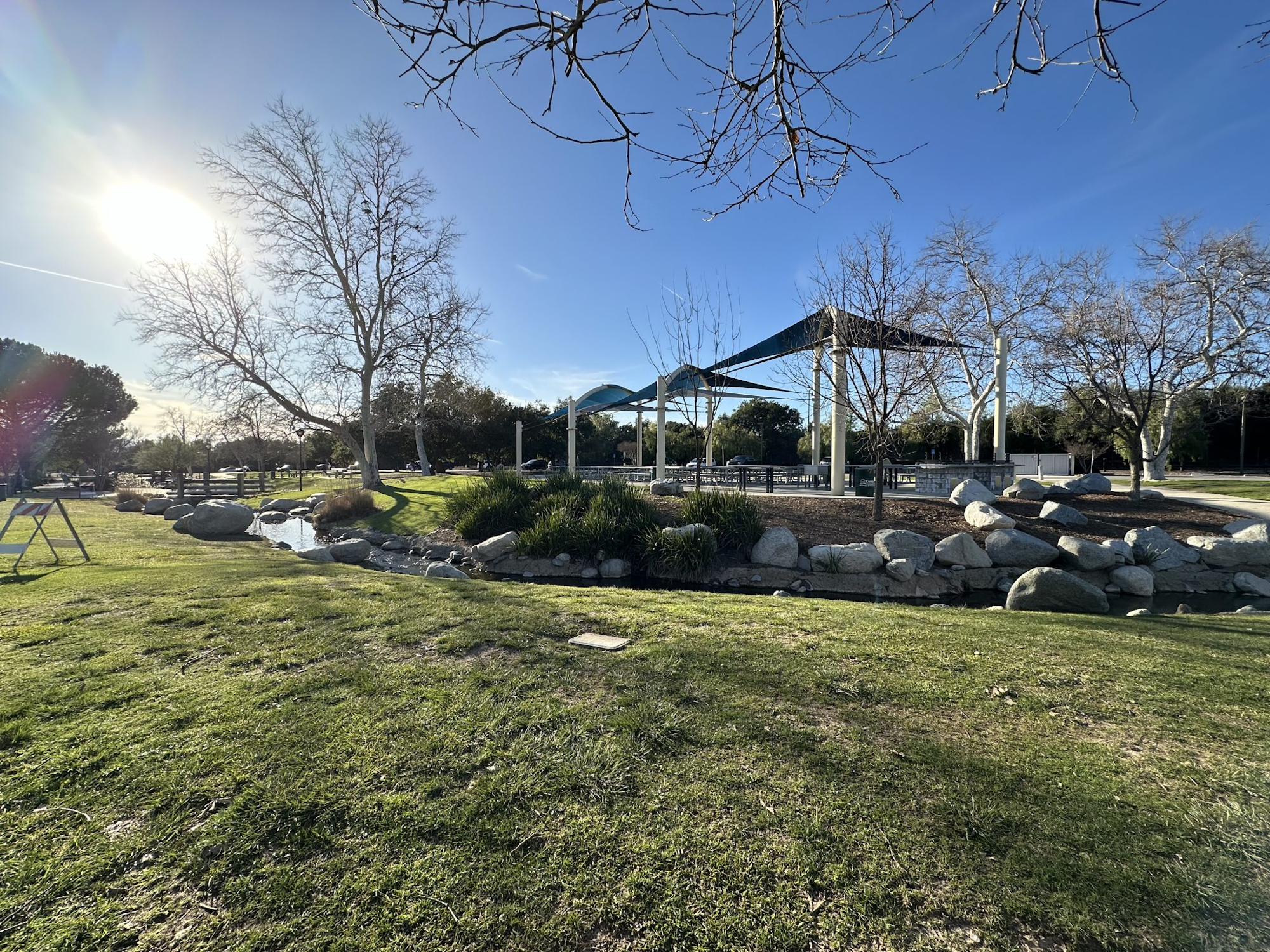 A sunny park scene with bare trees, a gazebo structure, rocks, and a small stream running through the grass.