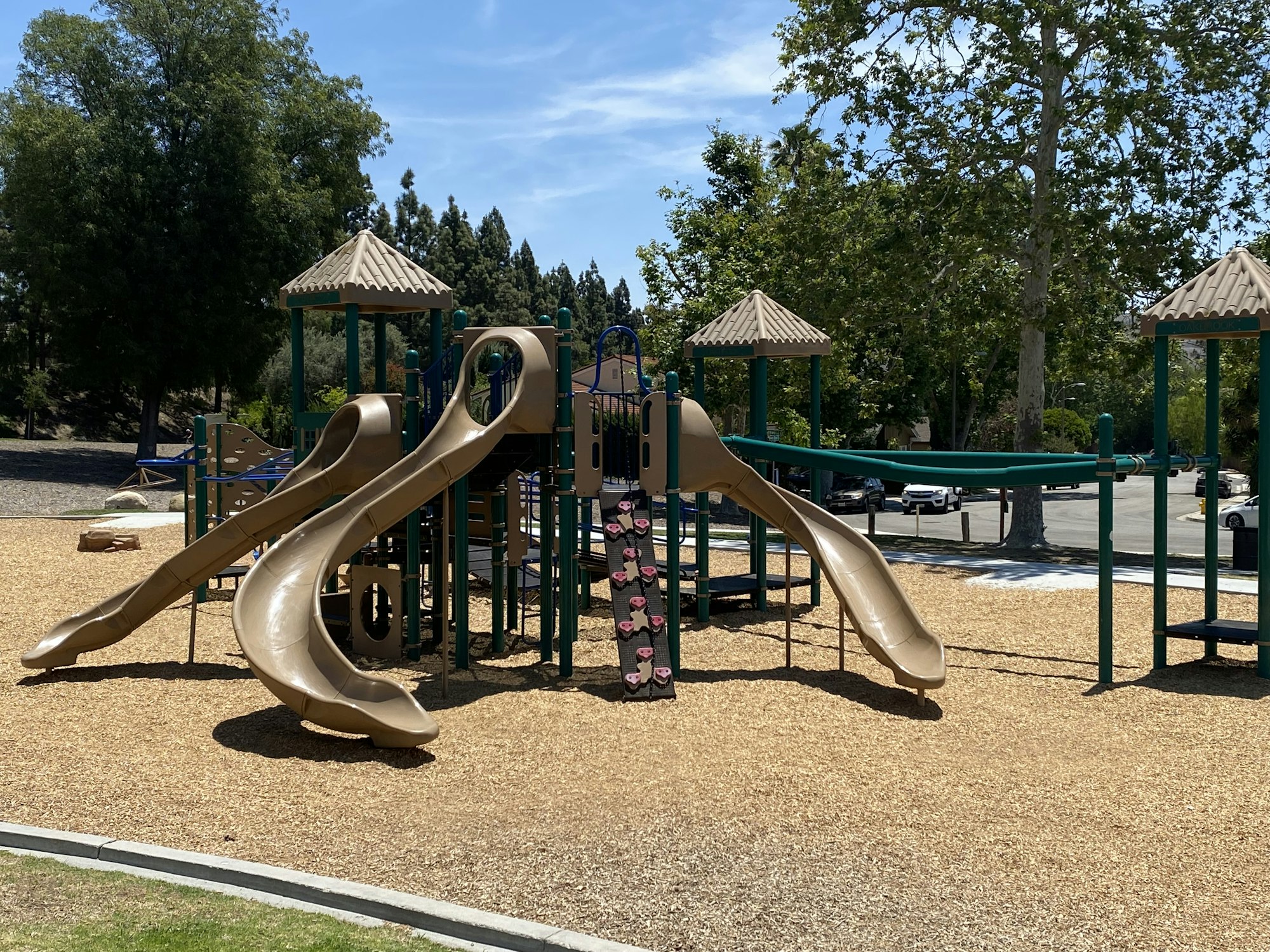 Playground with slides, climbing structures, and a woodchip surface, surrounded by trees and a parking area.