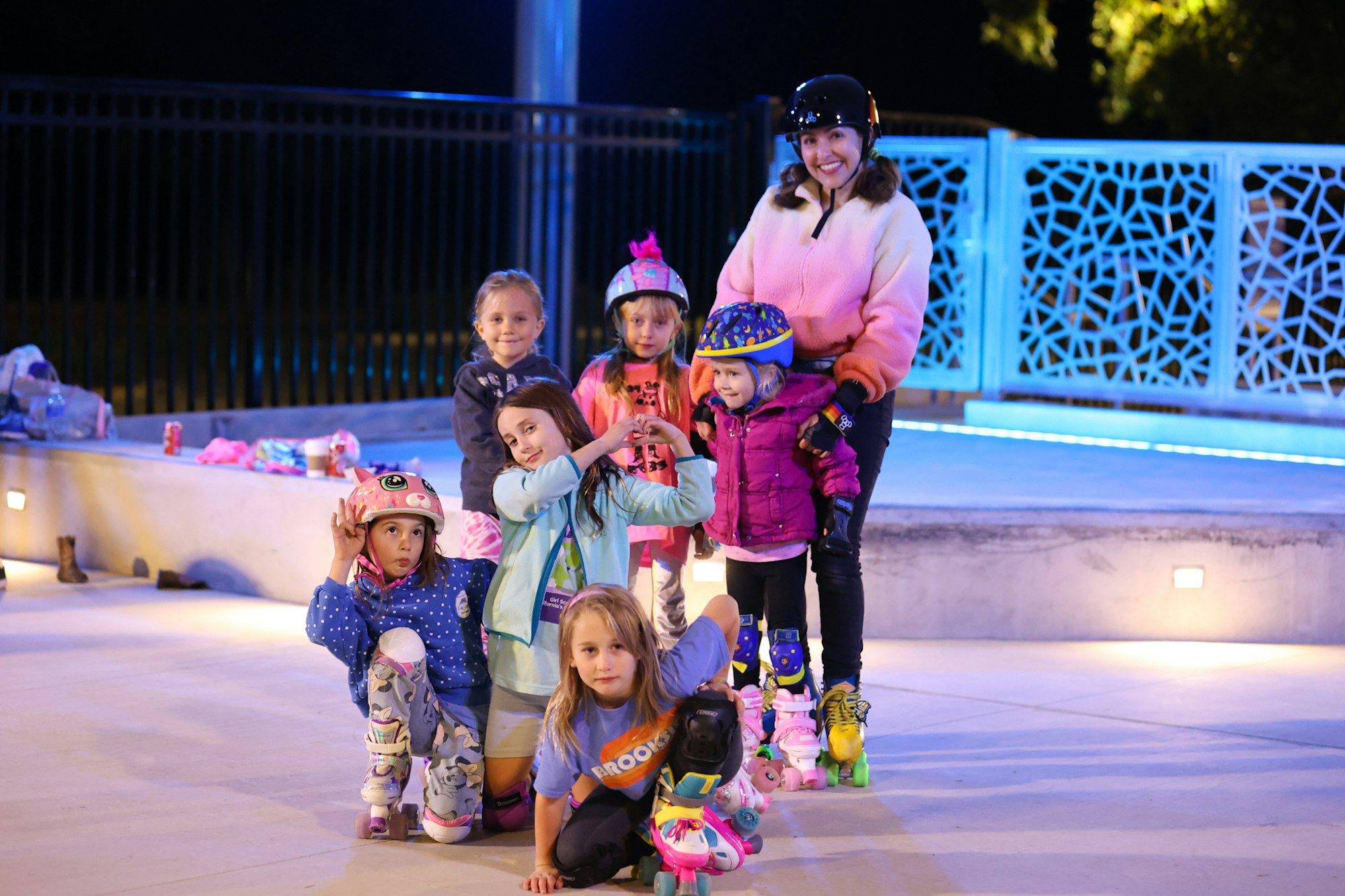 A group of children and a woman in roller skates pose happily together at night, with colorful outfits and helmets.