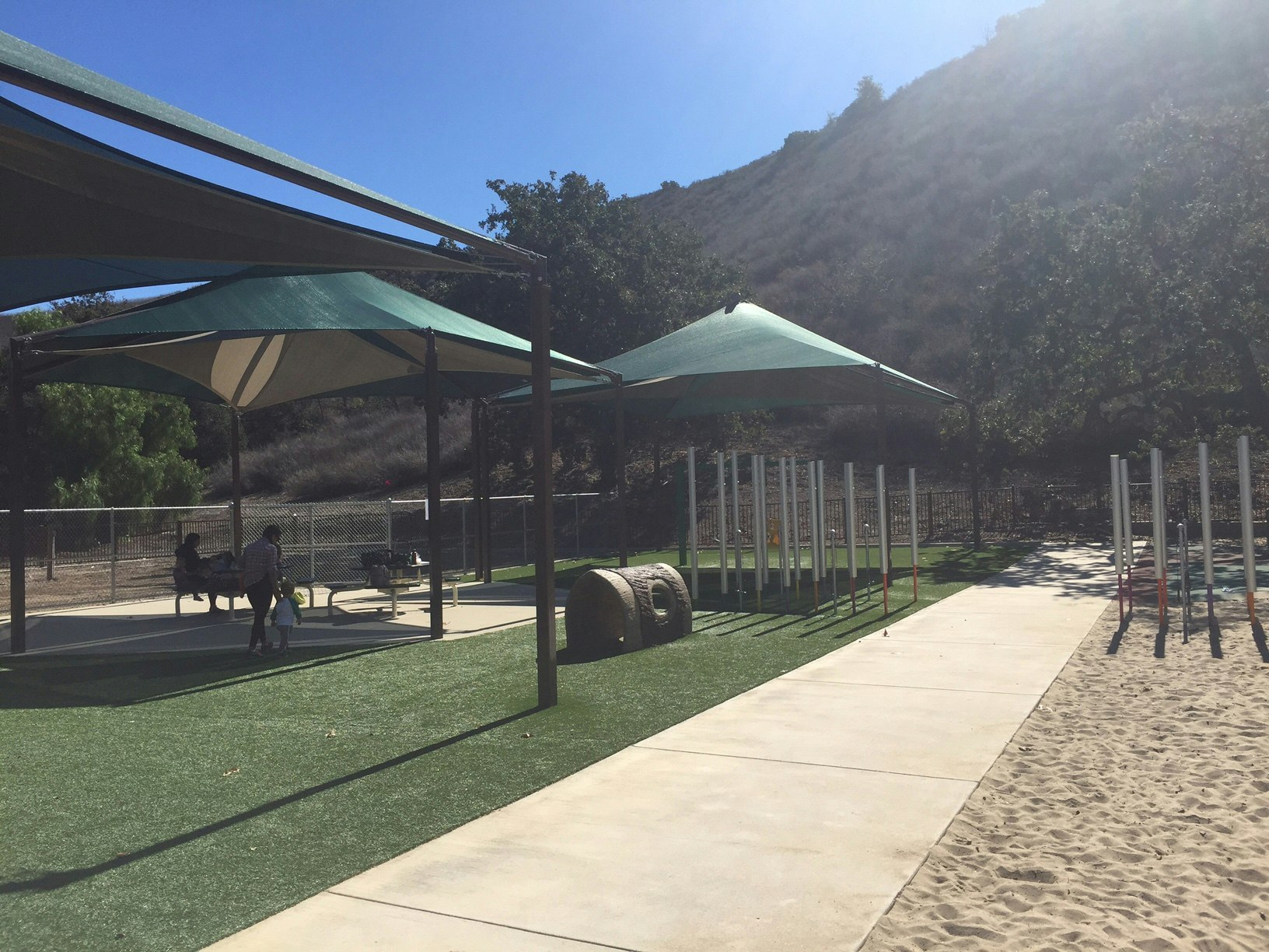 A playground with shade structures, artificial turf, benches, and play equipment; hills and trees in the background under a clear sky.
