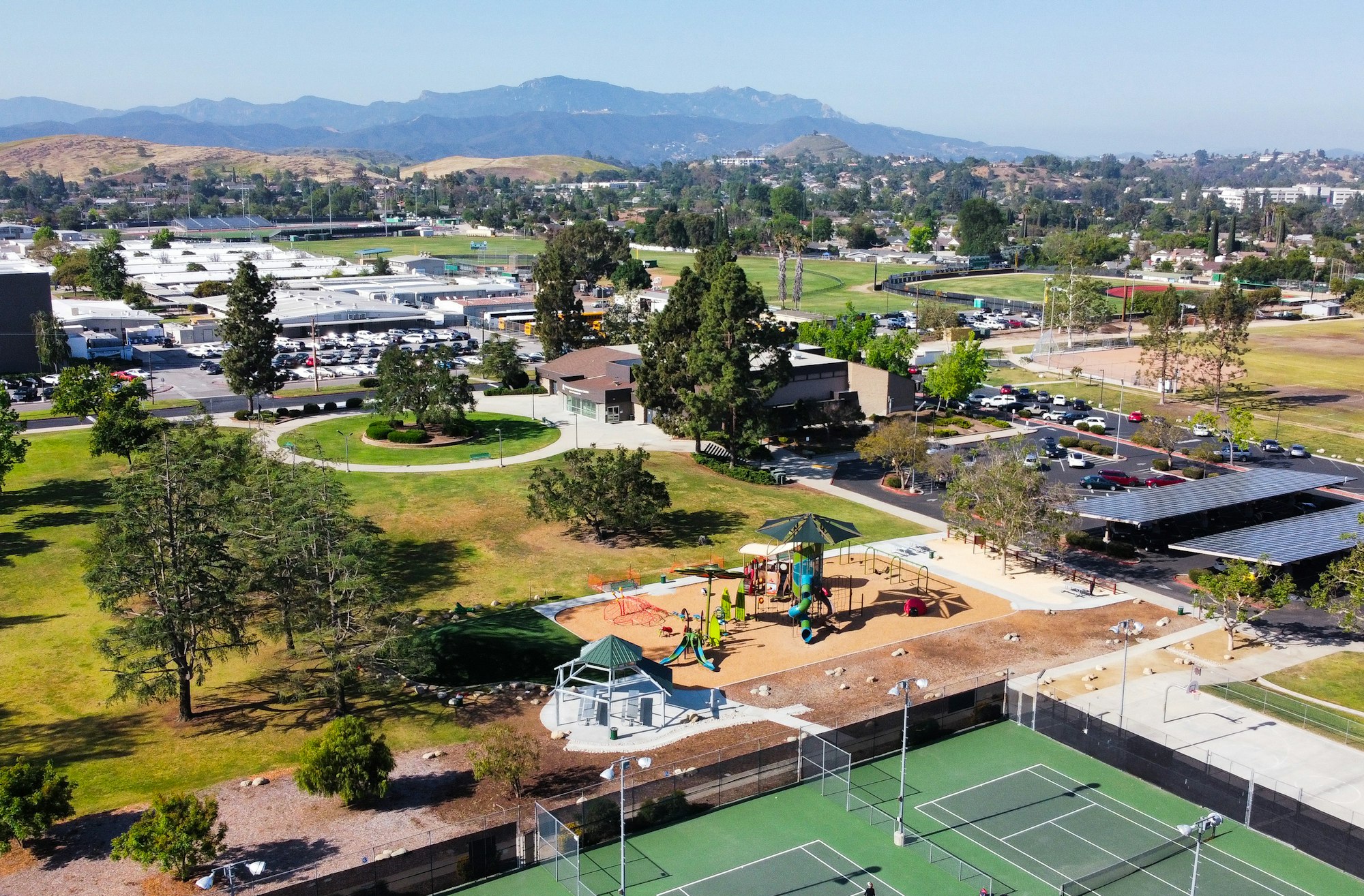 Aerial view of a school, playground, tennis courts, parking lot, and distant hills under a clear sky.